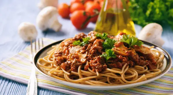 Une assiette de tagliatelles à la bolognaise, garnie de persil frais. Des champignons, tomates et une bouteille d'huile d'olive sont en arrière-plan. Une recette parfaite pour un repas réconfortant.