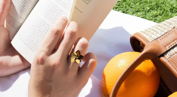 Mano con un anillo dorado con forma de flor, leyendo un libro junto a naranjas y un bolso de mimbre. Las tallas de anillos perfectas para cada ocasión.