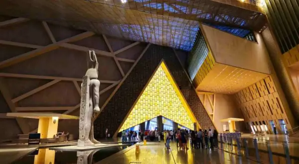 Wnętrze nowego muzeum w Kairze. Monumentalna rzeźba stoi przy basenie, a za nią znajduje się wejście w kształcie piramidy, oświetlone od środka.