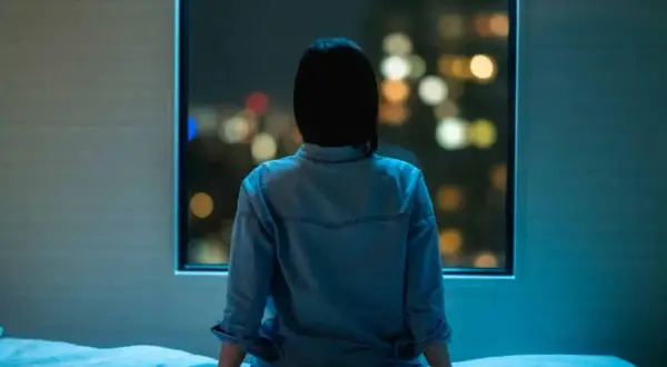 A person sits on a bed, looking out at city lights, feeling the weight of anxiety even when sleeping next to someone.