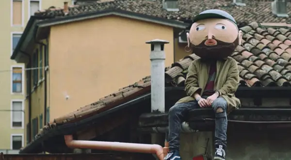 Un personaggio con una maschera gigante, simile a un cratere, siede sui tetti. I testi di Frah Quintale potrebbero raccontare la sua storia.