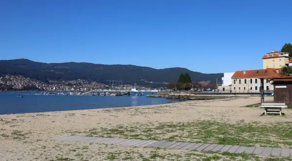La playa de arealonga, con arena dorada y vegetación dispersa, se extiende hacia un mar azul tranquilo. Al fondo, un puerto deportivo con barcos y una ciudad en la ladera de una montaña verde.