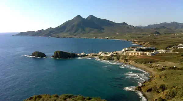 Vista panorámica de la cala del Toro, con sus aguas turquesas, rocas emergiendo y un pueblo blanco al fondo, bajo montañas verdes.