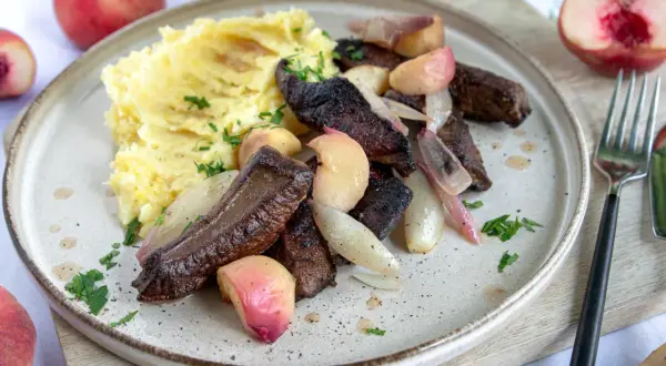 Leber braten ohne Mehl: zarte Leberstücke mit karamellisierten Zwiebeln und Pfirsichspalten, serviert mit cremigem Kartoffelpüree und frischer Petersilie.