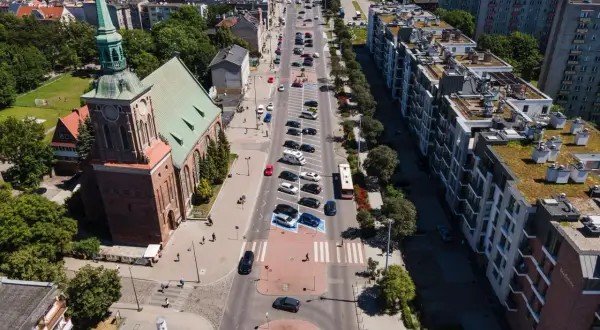 Widok z lotu ptaka na miasto z kościołem i długimi ogrodami. Ulicą jedzie autobus, a obok parkują samochody.