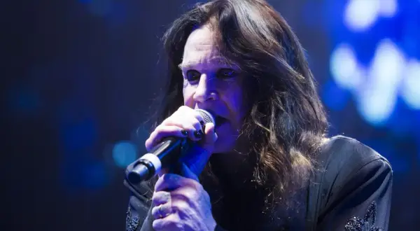 Ozzy Osbourne śpiewa do mikrofonu podczas koncertu, jego długie włosy opadają na twarz.