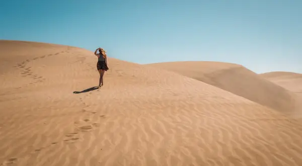 Kobieta spaceruje po piaszczystych wydmach Dunas de Maspalomas, zostawiając ślady na piasku pod błękitnym niebem.