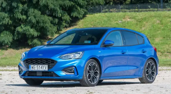 Niebieski Ford Focus 1.0 EcoBoost z czarnym dachem, zaparkowany na żwirowej nawierzchni, na tle zieleni.