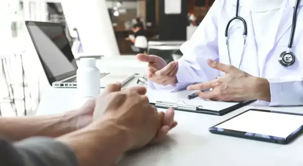 Lekarz omawia z pacjentem leczenie anty HCV. Na stole leżą tablet, laptop i butelka leków.