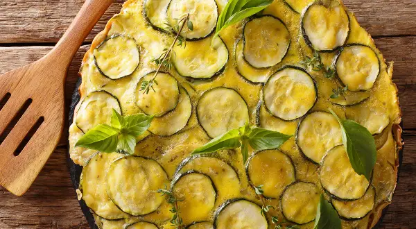 Une délicieuse scarpaccia de courgettes, garnie de basilic frais et de thym, prête à être servie avec une spatule en bois.