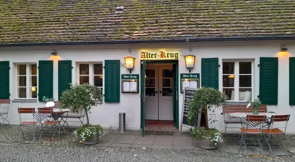 Der "Alter Krug" in Dahlem, ein charmantes Gasthaus mit grünen Fensterläden und einem moosbewachsenen Dach, lädt mit seinen Tischen im Freien zum Verweilen ein.