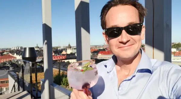 En man med solglasögon håller en rosa drink på en takterrass med utsikt över staden. En perfekt dag för en drink från arc blique by nobis.