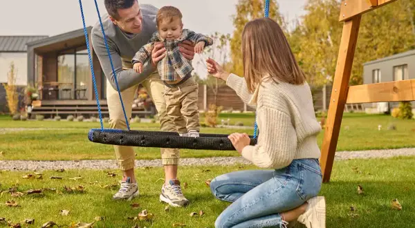 Rodzina cieszy się wspólnymi zabawami na podwórku. Tata trzyma dziecko na huśtawce, a mama zachęca do zabawy.