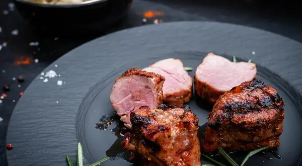Saftige schweine medaillons, perfekt gegrillt, liegen auf einem dunklen Teller, garniert mit Rosmarin. Daneben eine Schüssel mit cremiger Beilage.