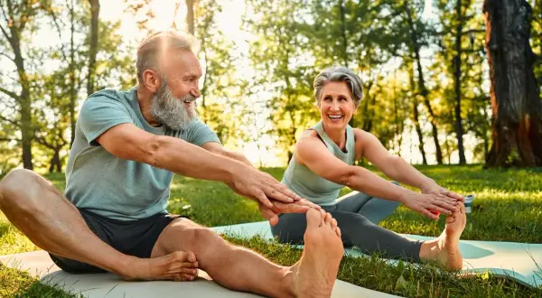 Couple souriant pratiquant le qi gong en plein air, étirant leurs jambes sur des tapis de yoga dans un parc ensoleillé.