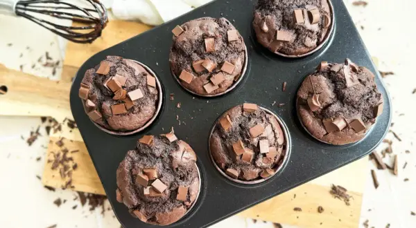 Sechs saftige, vegane Schokomuffins mit Schokostückchen in einer Muffinform. Ein Schneebesen mit Schokospuren liegt daneben.