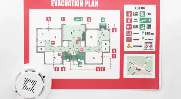 Piano di emergenza ed evacuazione con legenda, uscite, estintori e indicazioni. Un rilevatore di fumo è visibile in primo piano.