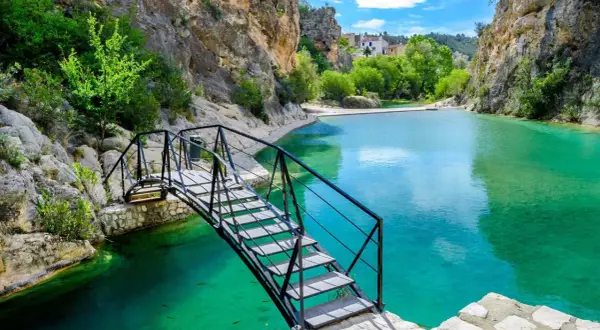 Puente metálico sobre aguas turquesas en uno de los lagos en Valencia, rodeado de rocas y vegetación.