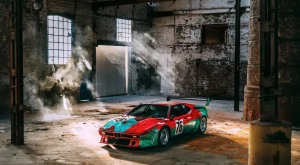 Un m1 coche de carreras con librea roja y verde, número 76, descansa en un almacén polvoriento con rayos de sol y humo.