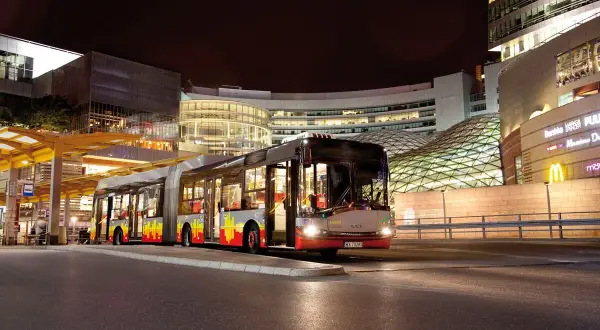Długi, przegubowy autobus linii 59 w Łodzi nocą, zatrzymuje się przy nowoczesnym centrum handlowym.