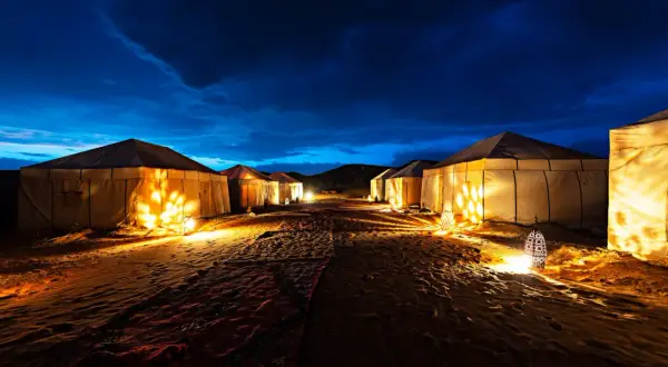 Campement de tentes illuminées dans le désert marocain, sous un ciel nocturne bleu profond. L'ambiance est magique.
