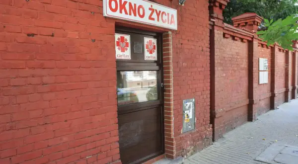 Czerwona ceglana ściana z napisem "OKNO ŻYCIA" i drzwiami. To miejsce daje szansę na nowe życie.