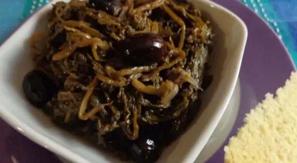Paparine verdura in umido con olive nere, servite in una ciotola bianca su un piatto viola, accanto a una fetta di pane.