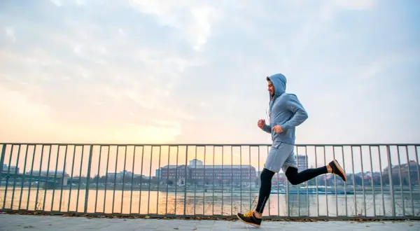 Mężczyzna w bluzie z kapturem uprawia jogging nad rzeką o zachodzie słońca. To świetny sposób na aktywność fizyczną.