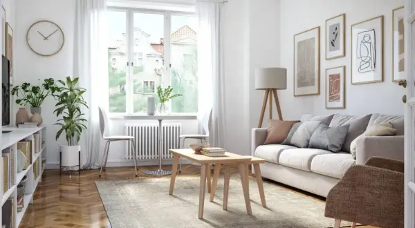 Ein helles Wohnzimmer mit Sofa, Couchtisch, Bücherregal und Pflanzen. Kunstwerke zieren die Wand, während durch das Fenster Tageslicht einfällt.