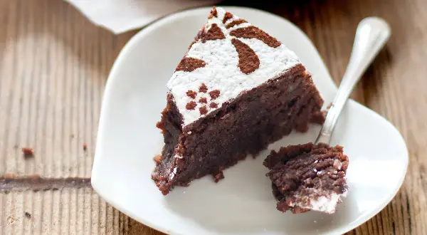 Una fetta di torta caprese ricetta originale napoletana, cosparsa di zucchero a velo e cacao, su un piatto bianco a forma di cuore.