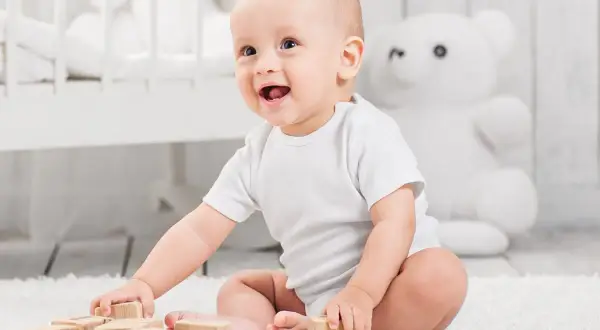 Uśmiechnięte niemowlę w białym body bawi się drewnianymi klockami na dywanie.