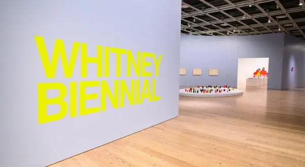 Bright yellow text announces the Whitney Biennial 2026. Inside the gallery, a circular display holds a colorful collection of small figures.
