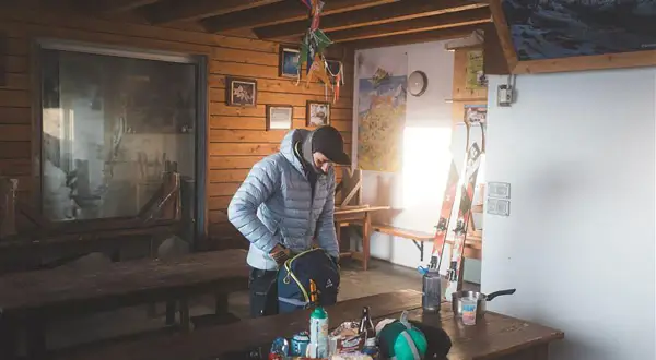 Dans une cabane de falaise, un skieur prépare son sac. Des skis, des cartes et des provisions sont visibles.