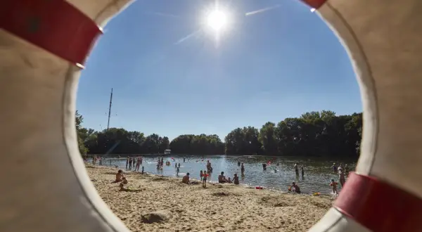 Sonniger Tag am Badesee in Hannover. Menschen planschen im Wasser und entspannen am Sandstrand, gesehen durch einen Rettungsring.