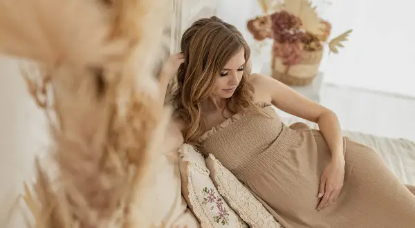 Piękna sesja ciążowa boho. Kobieta w beżowej sukience leży na łóżku, otoczona naturalnymi dekoracjami.