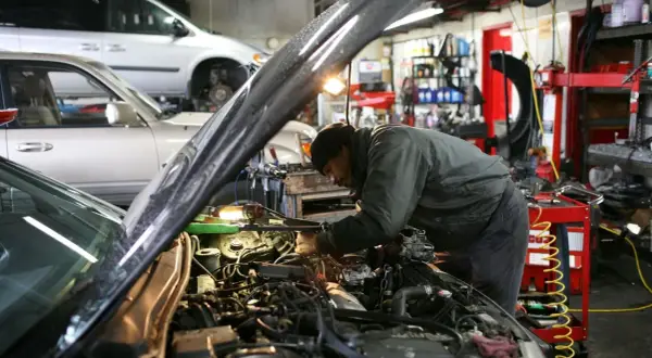 Mekaniker arbetar med en bilmotor i ett garage. En annan bil står i bakgrunden. Här erbjuds stor service.