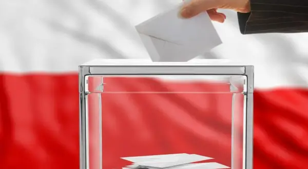 Dłoń wrzuca kartę do urny wyborczej. Polska flaga w tle symbolizuje kraj, od którego można głosować.