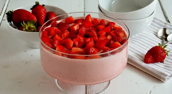 Delizioso mousse di fragole in una coppa di vetro, guarnito con fragole fresche a pezzetti. Accanto, una ciotola di fragole e ciotole bianche.