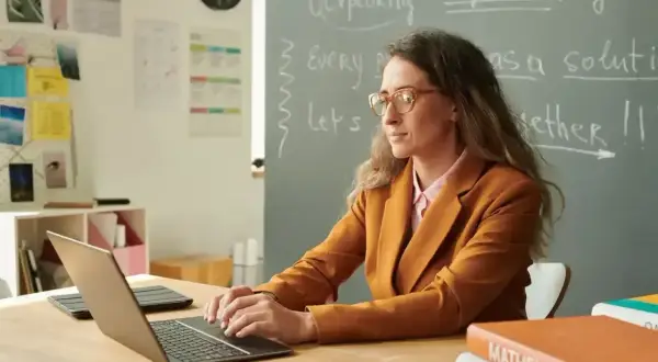 Nauczycielka pracuje przy laptopie. Na tablicy widnieje napis "English". Karta oceny pracy nauczyciela może być wypełniana cyfrowo.