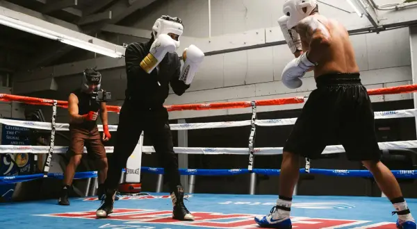 Two boxers in a brawler boxing style spar in a gym, one with white gloves and gold accents, the other shirtless. A third person films them.