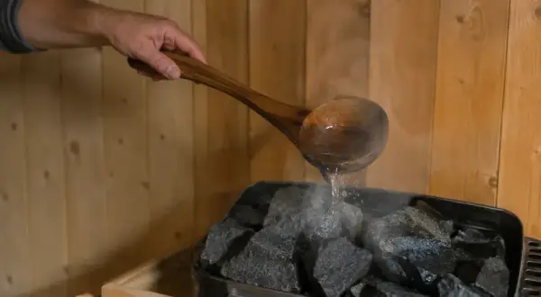 Ein Holzlöffel mit Wasser wird über heiße Steine gegossen, um Dampf zu erzeugen. So kann man sich den Duft von saunahonig selber machen.