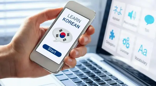 Dłoń trzyma smartfon z napisem "LEARN KOREAN" i flagą Korei. Obok laptop z ikonami do nauki języka koreańskiego od podstaw.