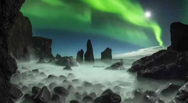 Grüne Nordlichter tanzen über einer felsigen Küste. Ein magischer Septemberabend auf einer Insel, wo das Meer in Nebel gehüllt ist.