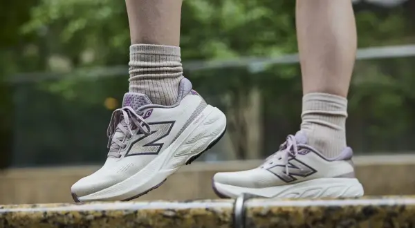Scarpe da tennis New Balance in beige e viola, con calzini beige arricciati, su uno sfondo verde sfocato.