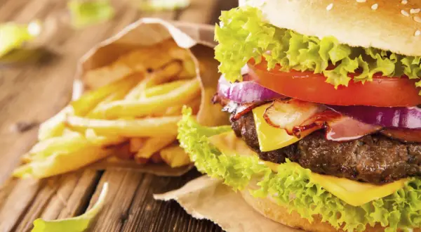 A close-up of a gourmet hamburger with lettuce, tomato, onion, bacon, and cheese, next to a bag of french fries.