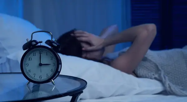 Budzik pokazuje 3:00. Osoba w łóżku, z rękami na głowie, cierpi z powodu problemów ze snem.