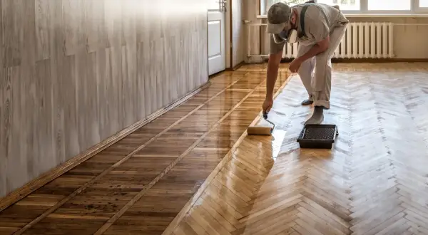 Un homme, masqué, utilise un rouleau pour vitrifier ou cirer un parquet en bois, lui redonnant son éclat.