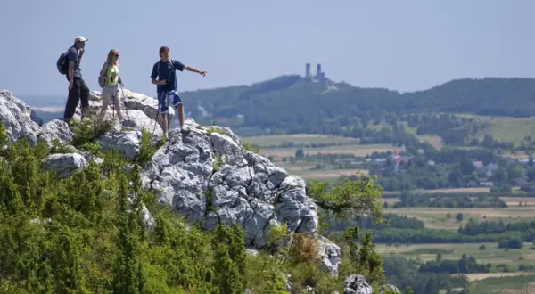 Trzech turystów na skale podziwia krajobraz Gór Świętokrzyskich. Jeden z nich wskazuje kierunek, gdzie w oddali widać ruiny zamku.