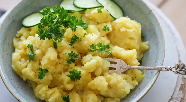 Schüssel mit bayrischem Kartoffelsalat, garniert mit Petersilie und Gurkenscheiben. Eine Gabel steckt darin.