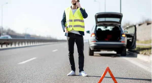 Mann mit Warnweste telefoniert neben seinem Auto mit offenem Kofferraum. Ein Warndreieck liegt auf der Straße. Wie viele Warnwesten im Auto sind, ist unklar.